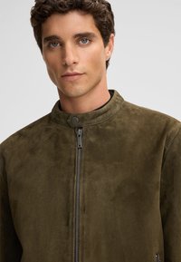 Olijfgroene suede jas met een opstaande kraag, ritssluiting aan de voorkant en een subtiele textuur. Minimalistisch ontwerp zonder zichtbare versieringen of patronen.