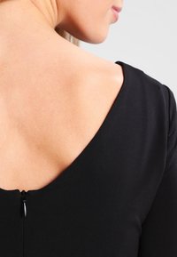 Černý top s hlubokým V-výstřihem, který má malý zip na zadní straně a hladkou, matnou texturu látky.