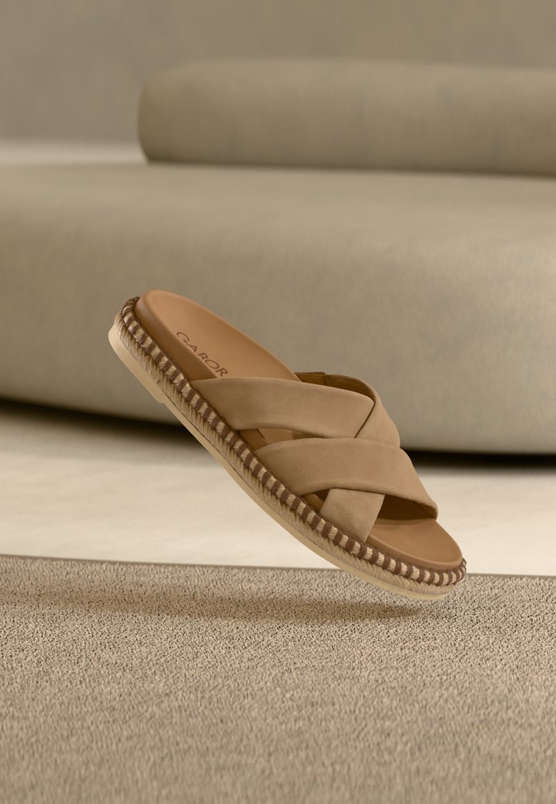 Sandale beige à enfiler avec des lanières croisées et des détails tissés sur la semelle, posée sur un tapis texturé dans une pièce aux tons neutres.