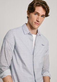 Ljusblå skjorta med knappar och blommönster, tillverkad av ett tyg med mjuk textur. Har en knappstängning och uppvikta ärmar.