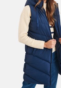 Gilet imbottito blu navy con cappuccio, caratterizzato da un design trapuntato, zip frontale completa e due tasche laterali. Indossato sopra un maglione color crema.
