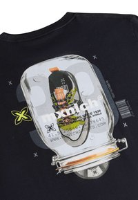 Černé tričko s grafikou skleněné nádoby, ve které je umístěná bota, s oranžovými a zelenými akcenty na botě a bílým textem, který detailně popisuje značku a původ.