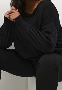 Pull noir texturé avec des manches larges côtelées, porté sur un pantalon noir. Le tissu a un léger éclat, ajoutant de la profondeur au design.