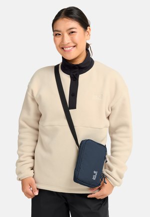 Lächende Person, die eine beige Fleecejacke mit schwarzem Kragen und schwarze Hose trägt, dazu eine marineblaue Umhängetasche mit weißem Logo.