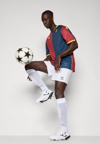 Rød og blå kortærmet trøje med gule detaljer, hvide shorts, hvide sokker og sort-hvide fodboldstøvler, som balancerer en mønstret fodbold.