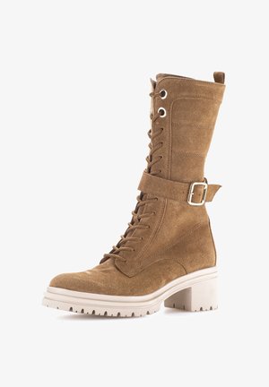 Tan suede mid-calf lace-up boot with white chunky sole and decorative buckle strap on side.
