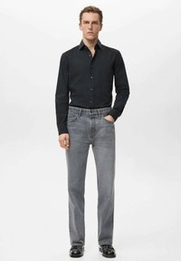 Czarna koszula z długim rękawem, rozpięta przy kołnierzyku, zestawiona z szarymi jeansami o prostych nogawkach. Dopełnieniem stroju są czarne buty o gładkiej fakturze.