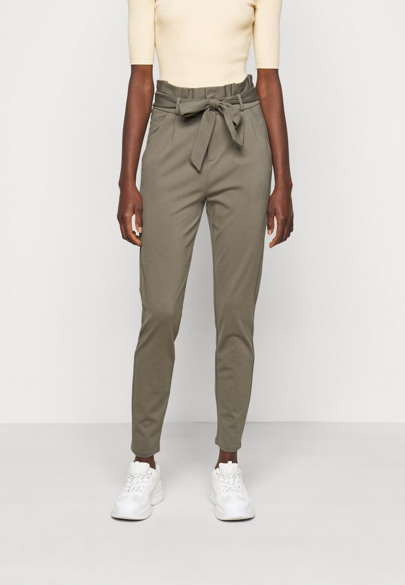 Femme portant un pantalon paperbag taille haute vert olive avec une ceinture en tissu nouée en nœud, un top côtelé crème et des baskets blanches.
