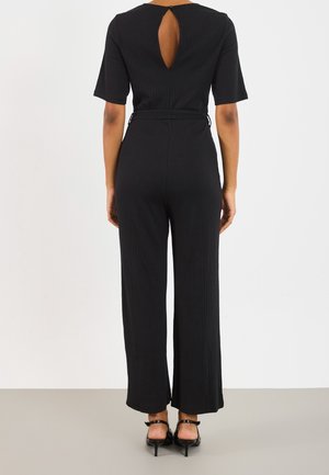 Femme debout de dos portant une combinaison noire à manches courtes avec une ouverture en forme de goutte dans le dos et des chaussures à talons noires.