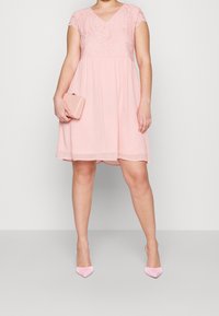 Kvinna iklädd en knälång rosa spetsklänning som håller en matchande rosa clutch, i sällskap av ljusrosa högklackade skor, stående mot en enkel bakgrund.