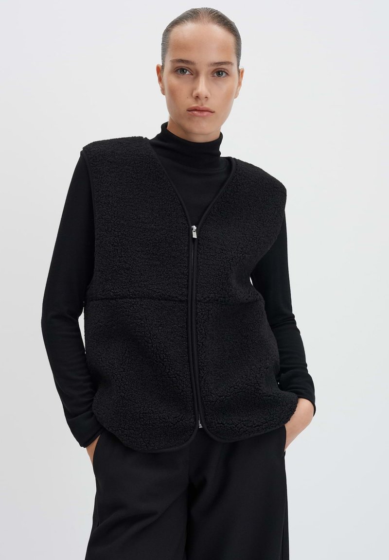 Gilet en polaire noir avec une fermeture éclair sur le devant, présentant un tissu texturé et un design sans manches, porté par-dessus un col roulé noir.