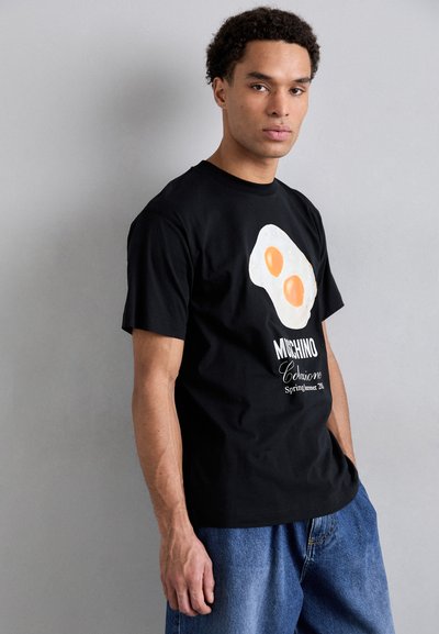 Black cotton T-shirt featuring a fried egg graphic in white and orange, with "Moschino" logo and "Spring/Summer 21" text at the bottom.