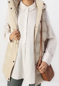 Personne portant un gilet matelassé beige par-dessus une chemise à rayures claires et boutonnée, tenant un sac en cuir marron en bandoulière par la sangle.