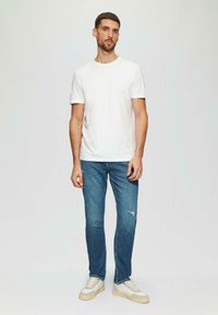 Witte katoenen t-shirt met ronde hals, blauwe denim jeans met verweerde details, en beige sneakers met grijze accenten, op een staande mannelijke model.