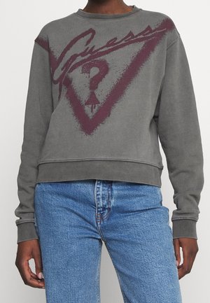 Persoon draagt een vervaagde grijze sweater met een bordeauxkleurig Guess-logo en een hooggetailleerde blauwe spijkerbroek, staand tegen een effen achtergrond.