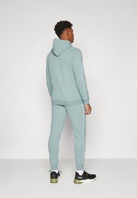 Haut à capuche bleu clair et jogging assorti avec des rayures blanches sur les côtés, fabriqués à partir de matériau doux. Des chaussures de sport noires complètent la tenue.