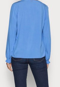 Person klädd i en långärmad blå blus med volangärmar och mörkblå denimjeans, sedd bakifrån.