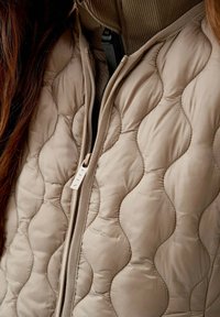 Gilet beige trapuntato con una superficie liscia, caratterizzato da cuciture ondulate e una zip frontale. Il colletto è a coste, aggiungendo profondità al design.