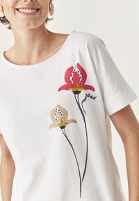 T-shirt en coton blanc avec un motif floral comprenant une fleur rouge et beige, des contours noirs et des accents verts. Col rond, manches courtes.