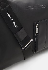 Bolso de cuero negro con una superficie texturizada, un bolsillo con cremallera y una correa con la marca "TOMMY JEANS" en blanco. Acentos en hardware plateado.