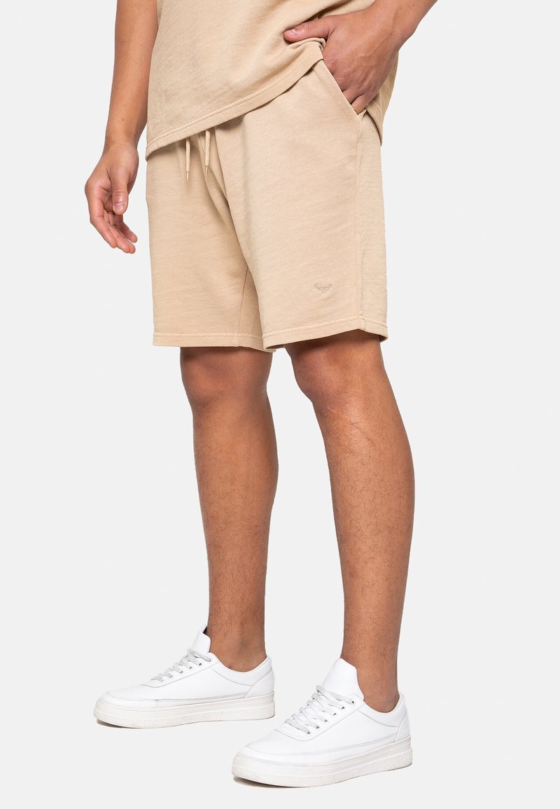 Shorts en coton beige avec une ceinture élastique et des poches latérales. Associés à des baskets blanches, ils présentent une texture lisse et un design décontracté.