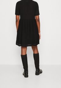 Vestido negro de manga corta con un corte holgado, de longitud por debajo de la rodilla, con un tejido fluido y una cintura fruncida. Combinado con botas negras hasta la rodilla.