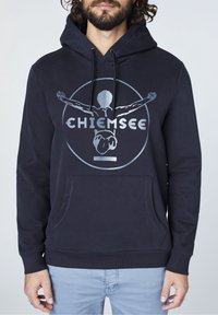 Mörkblå hoodie i bomullsblandning, med en framficka, dragkedjehuvud och en cirkulär silvergrafik med ordet "CHIEMSEE."