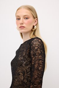 Top in pizzo nero con intricati motivi floreali, maniche lunghe e un colletto rotondo. La modella indossa grandi orecchini a cerchio dorati e ha i capelli biondi lisci.
