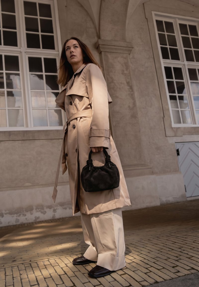 Femme portant un trench-coat beige et un pantalon large tient un petit sac à main noir, debout dans un bâtiment avec de grandes fenêtres à cadre blanc.
