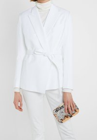 Vit blazer med bälte i midjan, slagna kragar och en enda ficka, kombinerad med vita byxor och en paljettclutch i silver och roséguld.