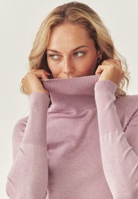Maglione a collo alto rosa chiaro con colletto e polsini a coste. Il tessuto sembra morbido e lavorato a maglia, caratterizzato da un design aderente.