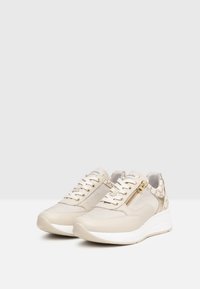 Baskets de sport beiges en matériaux mesh et cuir, avec une fermeture éclair sur le côté, des accents dorés et une semelle blanche épaisse.