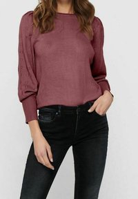 Maglione a maglia bordeaux con maniche aperte testurizzate, scollatura tonda e polsini a coste, abbinato a jeans neri aderenti.