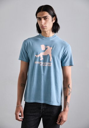 Light blue cotton t-shirt featuring a pink graphic design of a figure with a bucket, along with the text "KIDSUPER STUDIOS GET UR GET GOIN’" below.