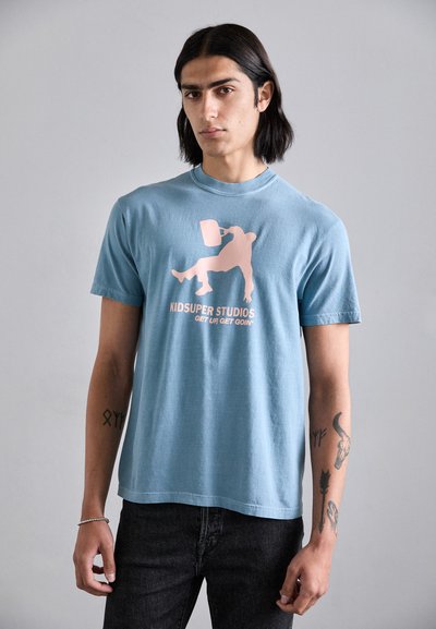 T-shirt en coton bleu clair présentant un design graphique rose d'une silhouette avec un seau, accompagné du texte "KIDSUPER STUDIOS GET UR GET GOIN’" en dessous.