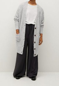 Hellgraue Strickjacke mit flauschiger Textur, offener Front, zwei seitlichen Taschen und braunen Knöpfen. Kombiniert mit schwarzen weiten Hosen und schwarzen Schuhen.