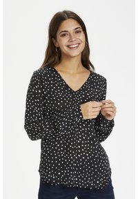 Schwarze Bluse mit weißen Polka-Punkten, langen Ärmeln, V-Ausschnitt, leichtem Stoff und gerafftem Detail vorne. Getragen mit dunkelblauen Jeans.