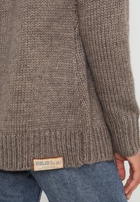 Brązowy sweter z dzianiny z prążkowanymi mankietami i dołem. Na dole po lewej stronie znajduje się metka z nazwą marki "Khujo." Tekstura jest gruba.