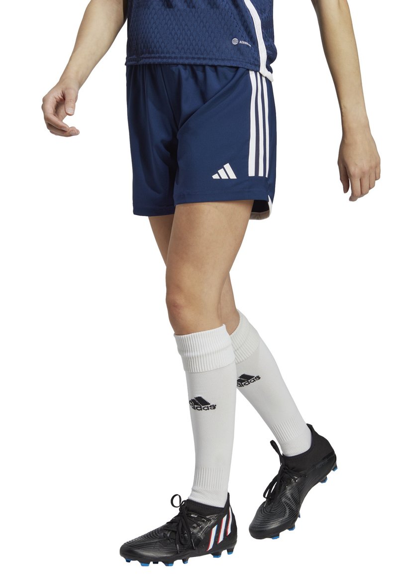 Marineblaue Sportshorts mit weißen Adidas-Streifen, kombiniert mit weißen kniehohen Socken mit schwarzem Logo und schwarzen Fußballschuhen.