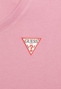Růžová pletenina s našitým trojúhelníkovým logem s nápisem "GUESS U.S.A." a červeným otazníkem uprostřed.