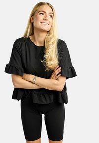 Schwarzes Oberteil mit Rüschenärmeln und gerafftem Saum, kombiniert mit schwarzen Radlerhosen. Das Material wirkt weich und figurbetont.