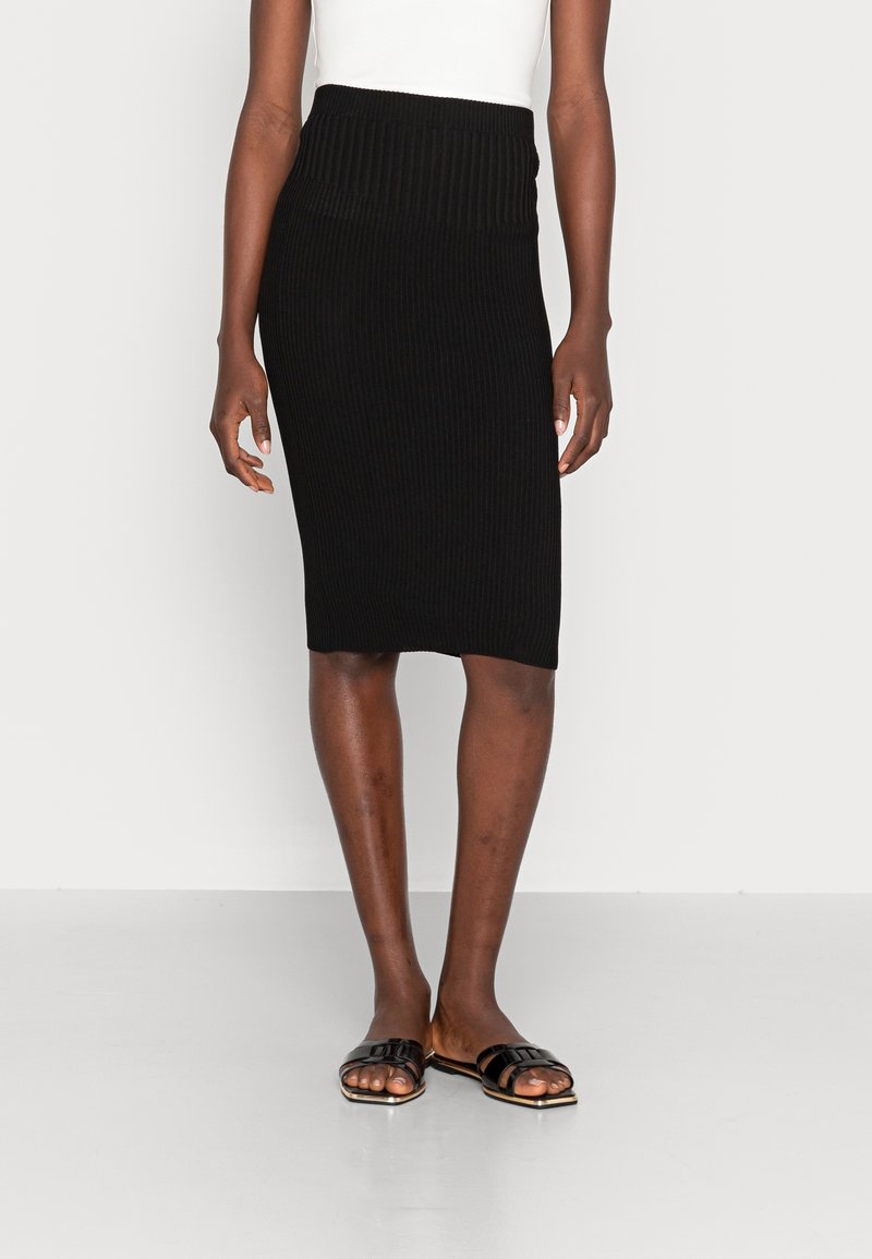 Jupe midi en tricot noir côtelé avec une taille haute. Portée avec des sandales plates noires. Le fond est blanc, mettant en valeur la tenue.