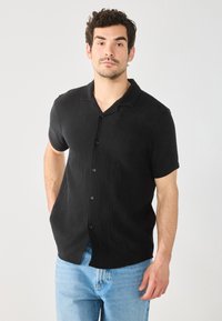 Camisa negra de manga corta con botones, con un patrón texturizado, cuello con solapa y botones negros, combinada con vaqueros azules claros.