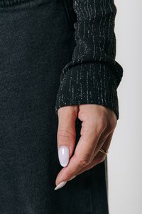 Top lung cu maneci lungi, de culoare neagră, cu dungi metalice verticale argintii, evidențiind detalii ale stofei texturate. O mână cu unghii roz pal susține marginea pantalonilor.