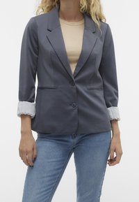 Šedý oblekový blazer se strukturovanými rameny, dvěma knoflíky a vyhrnutými světle šedými manžetami. Nosený přes béžový top a modré džíny.