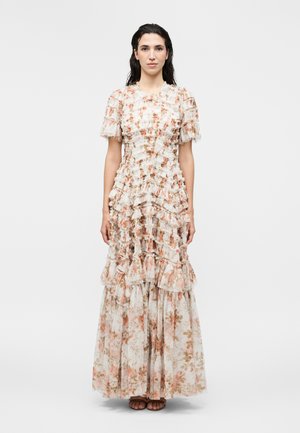 Kobieta w długiej, marszczonej sukience w kwiaty z krótkimi rękawami, stojąca boso na jednolitym białym tle.