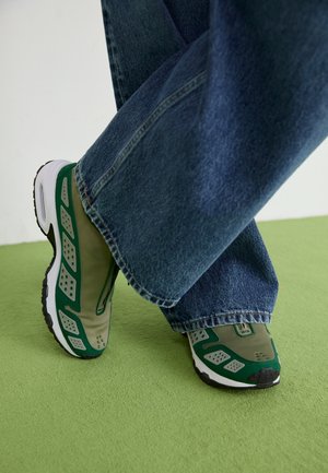 Personne portant des baskets vertes et grises avec des semelles blanches et un jean bleu debout sur un sol recouvert d'un tapis vert.