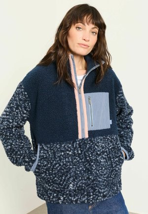 Frau trägt eine blau-navy strukturierte Fleecjacke mit Frontreißverschluss und Brusttasche, die Hände in den Taschen, steht vor einem schlichten Hintergrund.