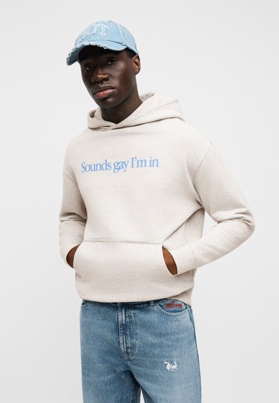 Bež duks s kapuljačom s plavim izvezenim natpisom "Sounds gay I'm in," upareno sa svijetloplavim trapericama koje na džepu imaju dizajn zeca.