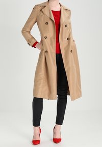 Beige trenchcoat met dubbele knoopsluiting, brede revers en zijzakken, gecombineerd met rode hakken en een rode top. Zwarte broek.